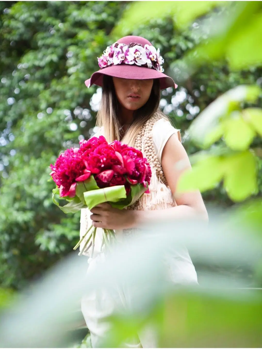 Mannequin debout au Jardin des Plantes à Paris, portant un chapeau prune décoré d'orchidées et tenant un grand bouquet de pivoines fuchsia de Cindy Gunther